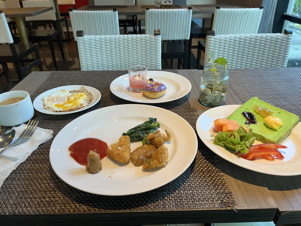 ホテルの朝食。目玉焼き、揚げ物、青菜、緑色の食パン、フルーツ、ドーナツなどを取り分けた皿がテーブルに並んでいる