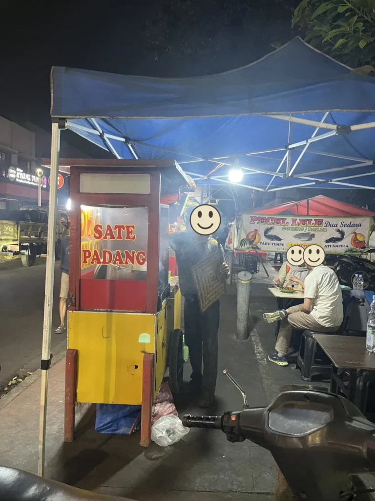 夜の歩道にある、赤と黄色のサテ・パダンの屋台。屋台の人がうちわを手に持っている